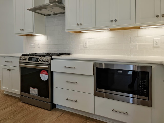 kitchen with appliances with stainless steel finishes, white cabinetry, decorative backsplash, light wood-type flooring, and wall chimney exhaust hood