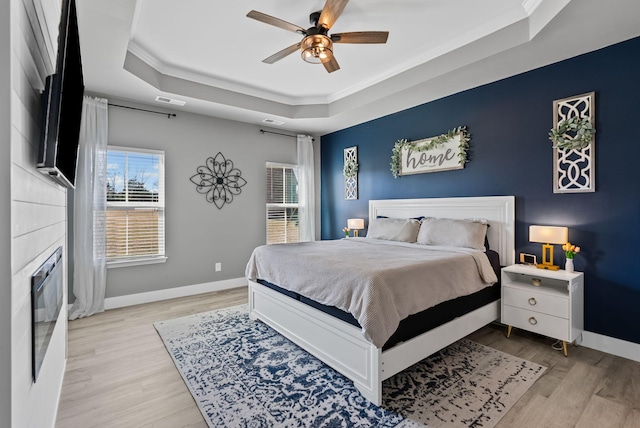 bedroom featuring a tiled fireplace, ceiling fan, a tray ceiling, crown molding, and light hardwood / wood-style flooring