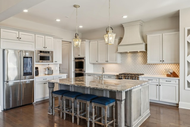 kitchen with stone counters, white cabinetry, stainless steel appliances, custom range hood, and a center island with sink