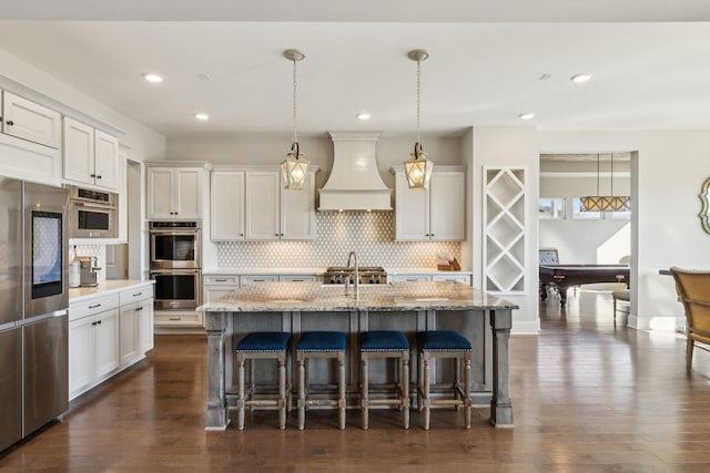 kitchen with appliances with stainless steel finishes, custom range hood, pendant lighting, and a kitchen island with sink