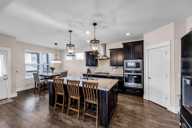 kitchen with pendant lighting, a breakfast bar area, stainless steel appliances, a center island with sink, and wall chimney exhaust hood