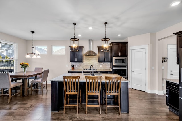 kitchen featuring wall chimney range hood, backsplash, stainless steel appliances, an island with sink, and decorative light fixtures