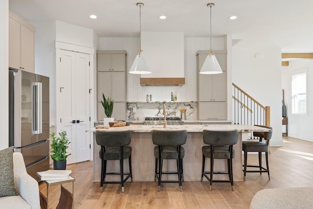 kitchen with high end fridge, custom range hood, decorative backsplash, a center island with sink, and decorative light fixtures