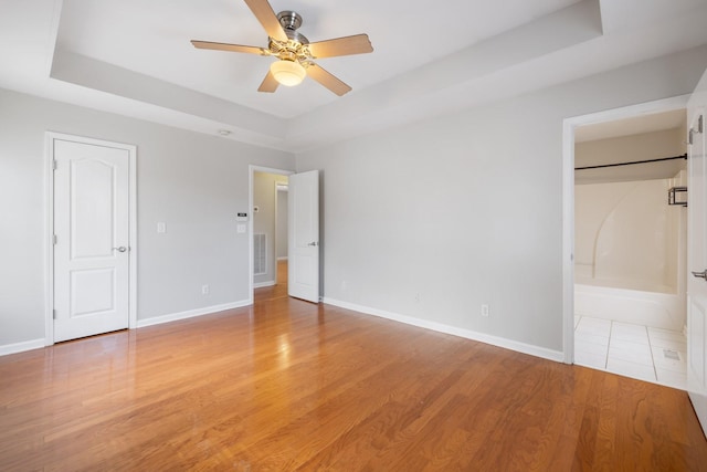 unfurnished bedroom featuring ceiling fan, a tray ceiling, a barn door, ensuite bath, and light hardwood / wood-style flooring