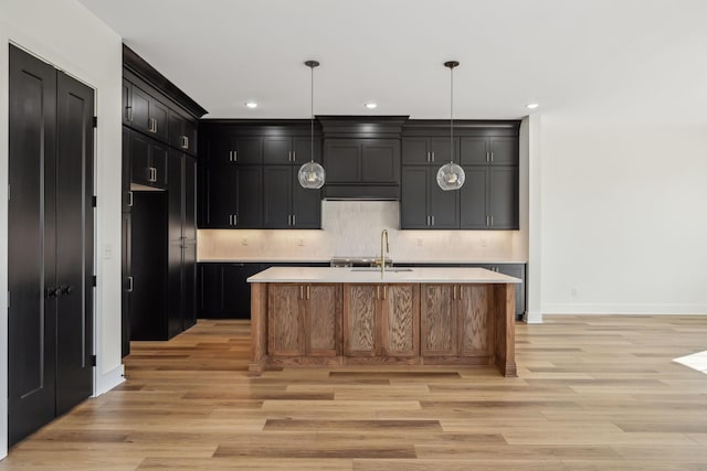 kitchen featuring sink, light hardwood / wood-style flooring, pendant lighting, a kitchen island with sink, and backsplash