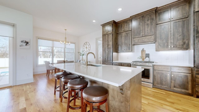 kitchen with decorative light fixtures, backsplash, a center island with sink, stainless steel gas range, and light hardwood / wood-style flooring