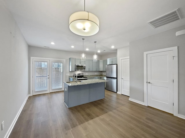 kitchen with gray cabinetry, hanging light fixtures, a center island with sink, appliances with stainless steel finishes, and light stone countertops