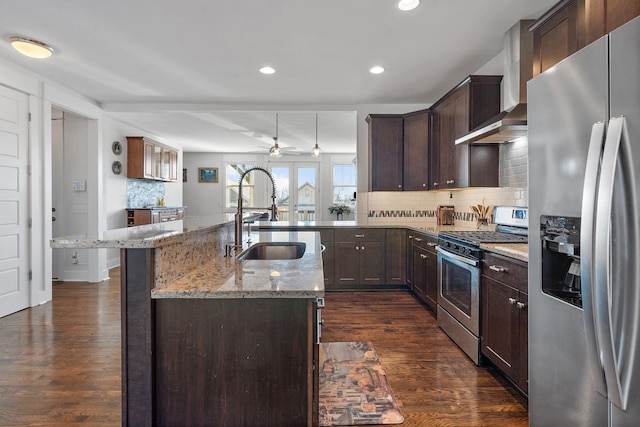 kitchen with wall chimney range hood, sink, a kitchen island with sink, stainless steel appliances, and light stone counters