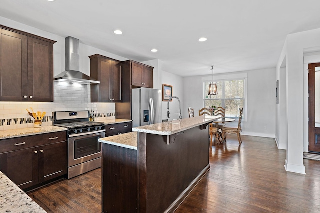 kitchen featuring pendant lighting, an island with sink, stainless steel appliances, light stone countertops, and wall chimney exhaust hood