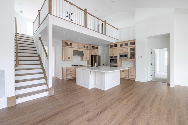 kitchen with wall chimney exhaust hood, a center island with sink, light hardwood / wood-style flooring, appliances with stainless steel finishes, and a towering ceiling