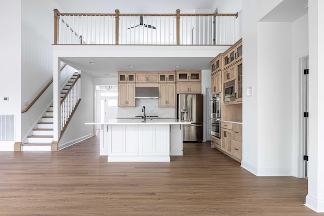 kitchen featuring wood-type flooring, an island with sink, stainless steel appliances, decorative backsplash, and wall chimney range hood