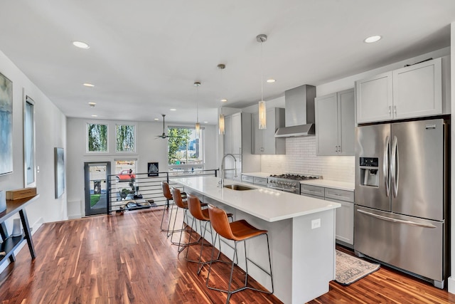 kitchen featuring wall chimney exhaust hood, sink, decorative light fixtures, appliances with stainless steel finishes, and an island with sink