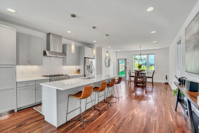 kitchen featuring pendant lighting, wall chimney range hood, dark wood-type flooring, stainless steel appliances, and an island with sink