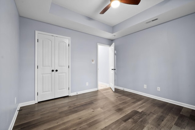 unfurnished bedroom with ceiling fan, a tray ceiling, dark hardwood / wood-style flooring, and a closet