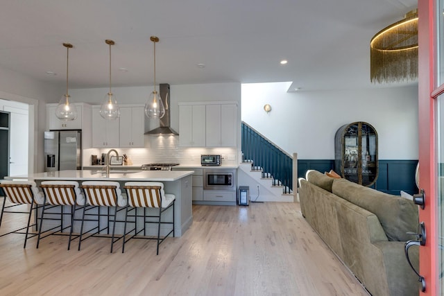 kitchen featuring wall chimney exhaust hood, white cabinetry, hanging light fixtures, appliances with stainless steel finishes, and a kitchen island with sink