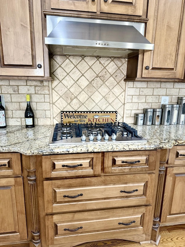 kitchen featuring wall chimney range hood, backsplash, light stone countertops, and stainless steel gas cooktop