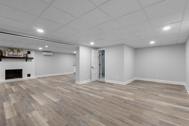 basement with a paneled ceiling, a brick fireplace, a wall mounted AC, and light hardwood / wood-style flooring