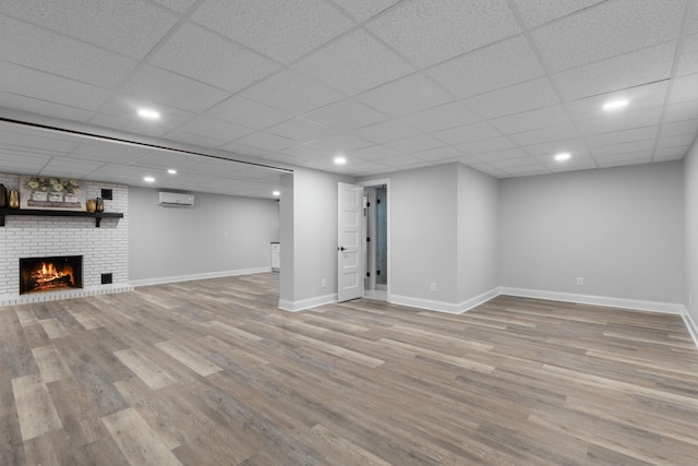 basement with a brick fireplace, a paneled ceiling, a wall unit AC, and light hardwood / wood-style floors