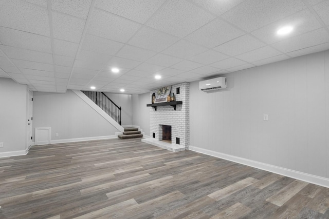 basement with a brick fireplace, wood-type flooring, a wall unit AC, and a paneled ceiling