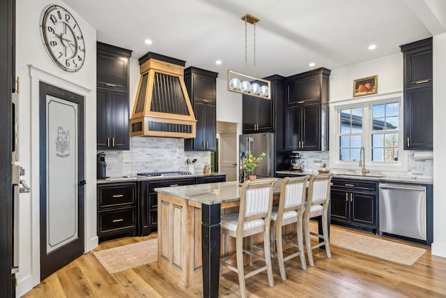 kitchen with sink, appliances with stainless steel finishes, an island with sink, decorative light fixtures, and custom exhaust hood