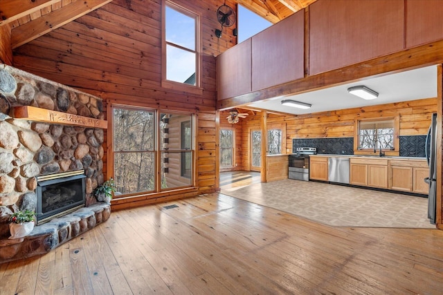 unfurnished living room featuring sink, light hardwood / wood-style flooring, high vaulted ceiling, a stone fireplace, and wood walls