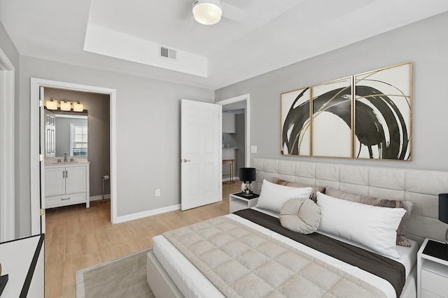 bedroom with connected bathroom, sink, a tray ceiling, and light wood-type flooring