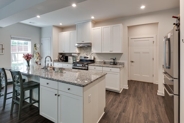 kitchen with appliances with stainless steel finishes, a kitchen island with sink, light stone countertops, white cabinets, and dark hardwood / wood-style flooring