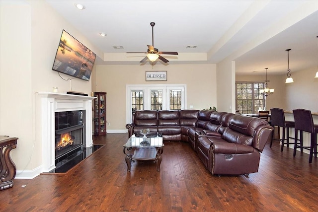 living room featuring a tray ceiling, dark wood-type flooring, a tile fireplace, and ceiling fan with notable chandelier