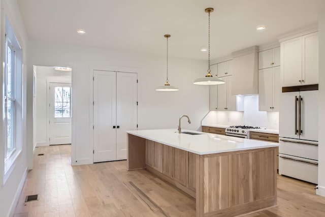 kitchen with high end white fridge, white cabinets, hanging light fixtures, a kitchen island with sink, and wall chimney range hood