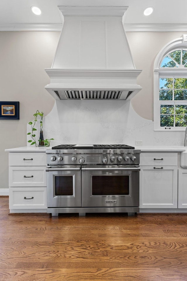 kitchen with custom exhaust hood, wood-type flooring, ornamental molding, range with two ovens, and white cabinets