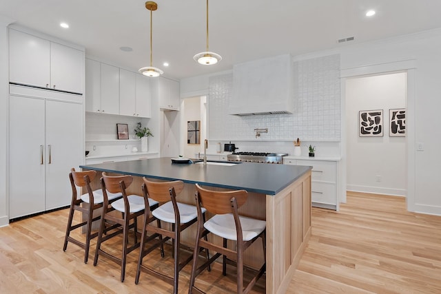 kitchen with premium range hood, sink, a center island with sink, light wood-type flooring, and white cabinets