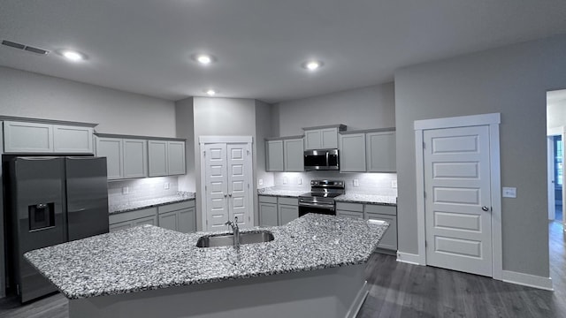 kitchen with dark wood-type flooring, sink, an island with sink, stainless steel appliances, and light stone countertops