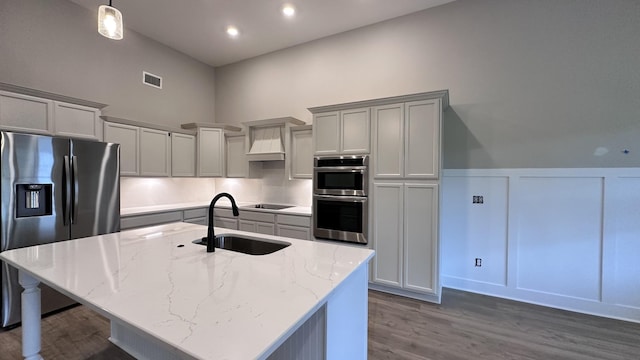 kitchen featuring premium range hood, appliances with stainless steel finishes, an island with sink, sink, and light stone counters