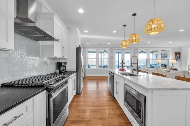 kitchen with wall chimney range hood, sink, appliances with stainless steel finishes, a kitchen island with sink, and decorative light fixtures