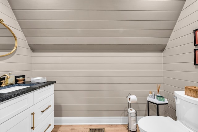 bathroom with hardwood / wood-style floors, wooden walls, vanity, vaulted ceiling, and toilet