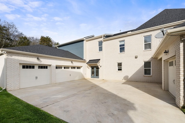 exterior space featuring a garage, brick siding, driveway, french doors, and roof with shingles