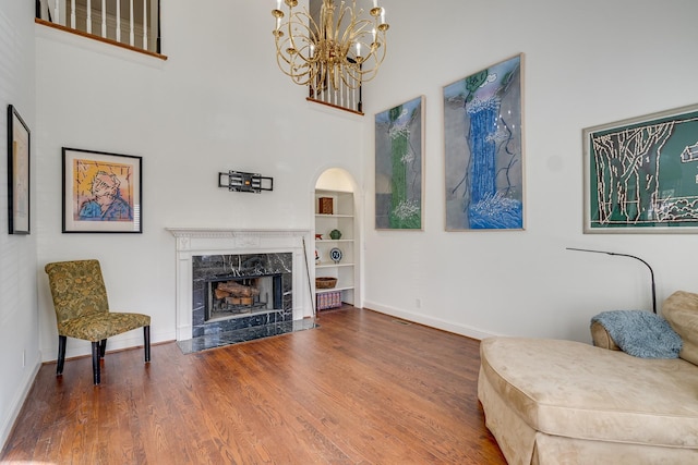 sitting room with a premium fireplace, a towering ceiling, wood-type flooring, built in shelves, and a chandelier