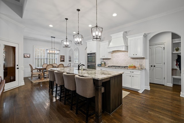 kitchen featuring appliances with stainless steel finishes, pendant lighting, white cabinets, a kitchen island with sink, and custom range hood