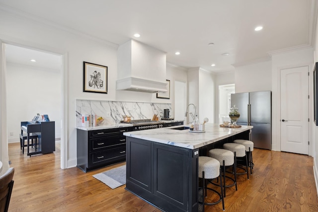 kitchen with sink, a center island with sink, custom range hood, stainless steel appliances, and light stone countertops