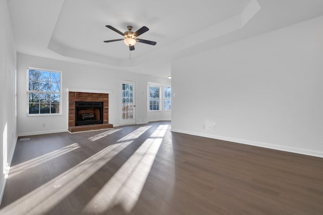 unfurnished living room featuring dark hardwood / wood-style flooring, a brick fireplace, a tray ceiling, and ceiling fan