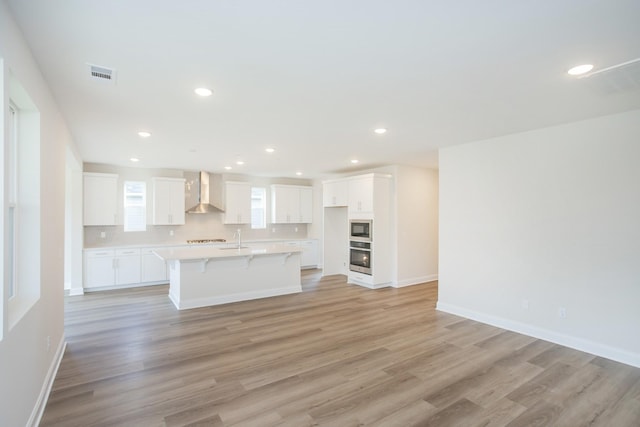 kitchen with wall chimney range hood, a kitchen island with sink, white cabinetry, oven, and light wood-type flooring