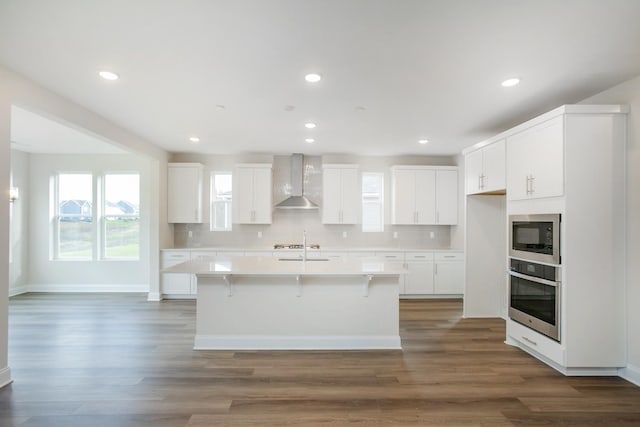 kitchen with white cabinetry, oven, wall chimney exhaust hood, and a center island with sink