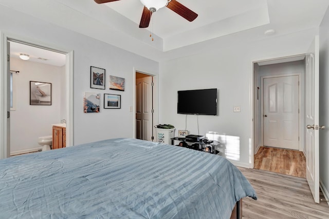 bedroom featuring connected bathroom, light hardwood / wood-style flooring, ceiling fan, and a tray ceiling