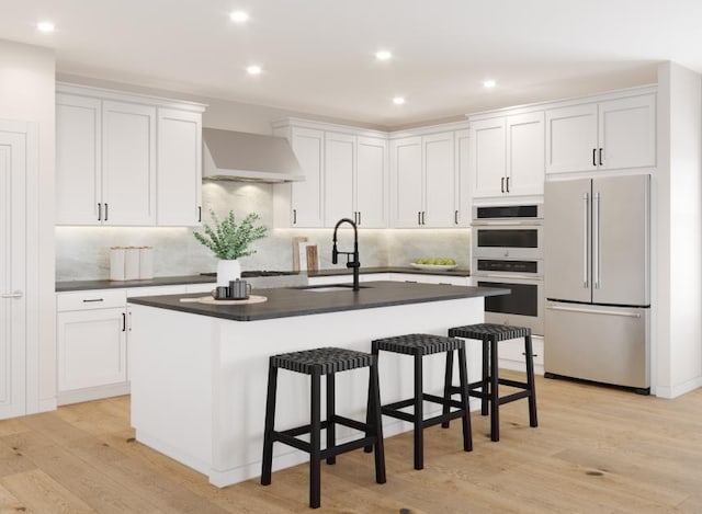 kitchen with wall chimney range hood, sink, appliances with stainless steel finishes, white cabinetry, and an island with sink