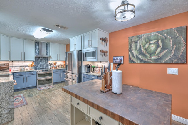 kitchen with butcher block countertops, wall chimney range hood, appliances with stainless steel finishes, backsplash, and blue cabinets