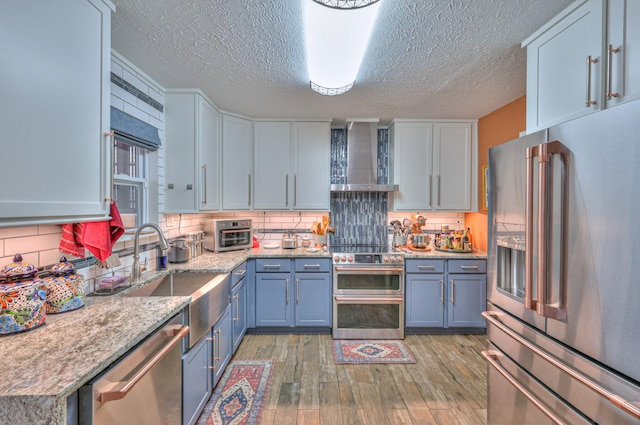 kitchen featuring backsplash, wall chimney exhaust hood, light stone counters, stainless steel appliances, and light hardwood / wood-style flooring