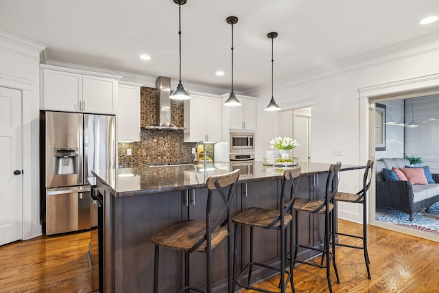 kitchen featuring white cabinetry, a center island with sink, dark stone countertops, appliances with stainless steel finishes, and wall chimney range hood