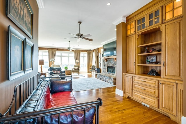 living room with ceiling fan, ornamental molding, a stone fireplace, and light wood-type flooring