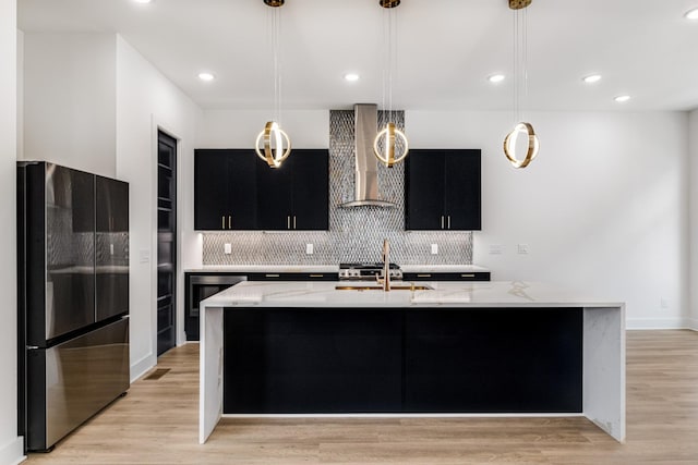 kitchen featuring pendant lighting, wall chimney range hood, stainless steel refrigerator, a kitchen island with sink, and light wood-type flooring