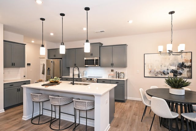 kitchen with gray cabinets, appliances with stainless steel finishes, an island with sink, sink, and hanging light fixtures